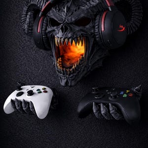 Puede incluir: Un soporte de pared de calavera demon&iacute;aca gris oscuro con un interior ardiente, que lleva auriculares negros y rojos. Dos mandos de juego, uno blanco y otro negro, son sostenidos por manos esquel&eacute;ticas debajo.