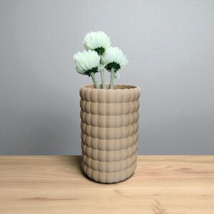 Pu&ograve; includere: Un vaso beige strutturato con un design arrotondato a forma di bolla, che contiene tre fiori bianchi con steli verdi. Il vaso &egrave; posto su una superficie di legno contro uno sfondo grigio neutro. I fiori aggiungono un tocco di freschezza.