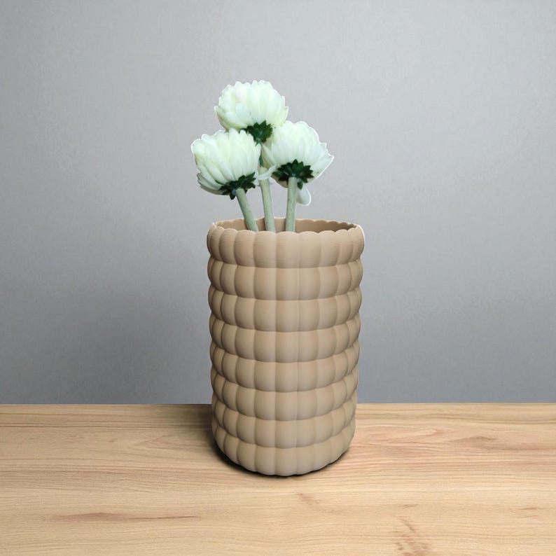 Pu&ograve; includere: Un vaso beige strutturato con un design arrotondato a forma di bolla, che contiene tre fiori bianchi con steli verdi. Il vaso &egrave; posto su una superficie di legno contro uno sfondo grigio neutro. I fiori aggiungono un tocco di freschezza.