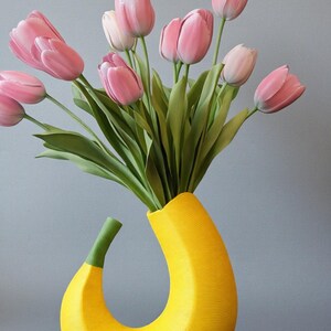 May include: A unique yellow vase shaped like a banana, holding a bouquet of pink tulips with green stems and leaves. The vase has a textured surface and a curved design, set against a gray background.
