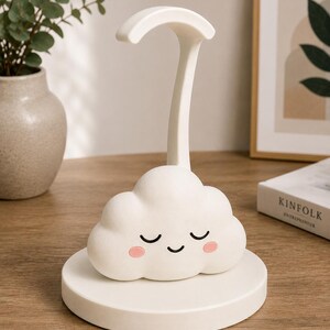 K&ouml;nnte beinhalten: Eine wei&szlig;e, wolkenf&ouml;rmige Schreibtischlampe mit einem l&auml;chelnden Gesicht, geschlossenen Augen und rosigen Wangen. Die Lampe hat eine gebogene Oberseite und einen hohen, schlanken Stiel und steht auf einem runden Sockel. F&uuml;r Schreibtisch oder Nachttisch.