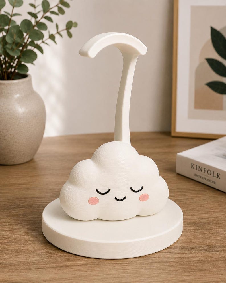 K&ouml;nnte beinhalten: Eine wei&szlig;e, wolkenf&ouml;rmige Schreibtischlampe mit einem l&auml;chelnden Gesicht, geschlossenen Augen und rosigen Wangen. Die Lampe hat eine gebogene Oberseite und einen hohen, schlanken Stiel und steht auf einem runden Sockel. F&uuml;r Schreibtisch oder Nachttisch.