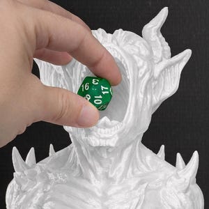 May include: A white, detailed sculpture of a monster with horns and spikes, holding its mouth open. A green, multi-sided die is held in front of the monster's mouth, with the number 3 visible. The sculpture is set against a black background.
