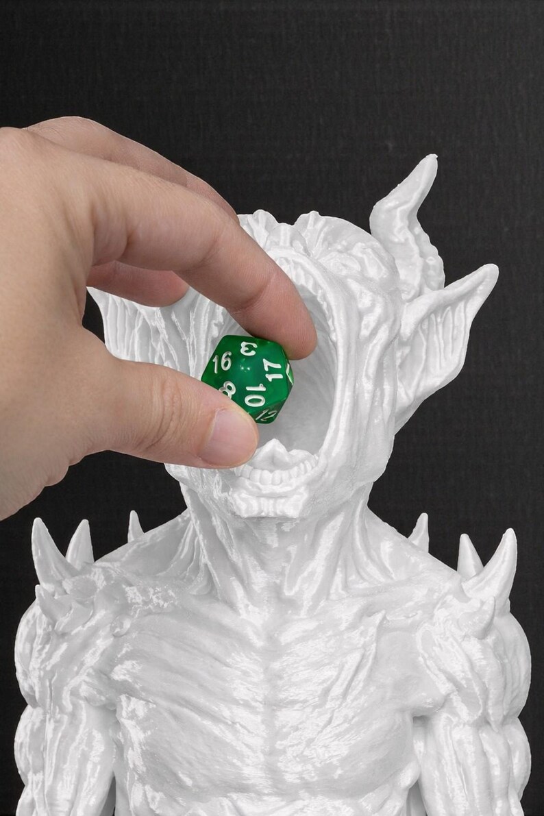 May include: A white, detailed sculpture of a monster with horns and spikes, holding its mouth open. A green, multi-sided die is held in front of the monster's mouth, with the number 3 visible. The sculpture is set against a black background.