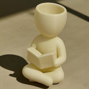 Puede incluir: Figura de color crema de una persona sentada con las piernas cruzadas y leyendo un libro. La figura tiene una cabeza grande y abierta, lo que sugiere una maceta o un jarr&oacute;n. El libro se sostiene en el regazo de la figura, y el dise&ntilde;o general es minimalista y moderno.