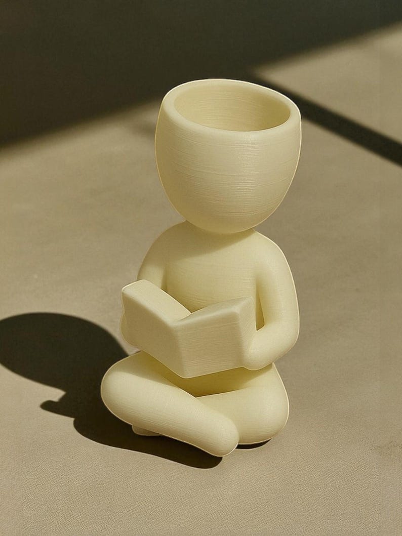 Puede incluir: Figura de color crema de una persona sentada con las piernas cruzadas y leyendo un libro. La figura tiene una cabeza grande y abierta, lo que sugiere una maceta o un jarr&oacute;n. El libro se sostiene en el regazo de la figura, y el dise&ntilde;o general es minimalista y moderno.