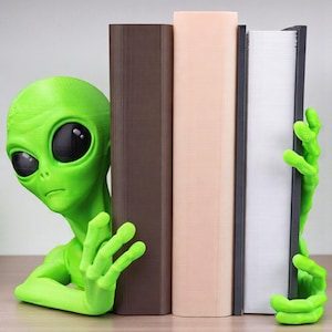 May include: A green alien-shaped bookend with large black eyes and slender fingers. The alien is peeking out from behind a row of books in shades of brown, beige, and white, creating a fun and unique bookshelf display.