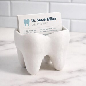May include: A white, tooth-shaped business card holder containing a stack of cards. The cards display the text "Dr. Sarah Miller DENTISTRY" and contact details. The holder is made from a smooth, solid material.