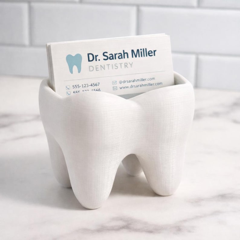 May include: A white, tooth-shaped business card holder containing a stack of cards. The cards display the text "Dr. Sarah Miller DENTISTRY" and contact details. The holder is made from a smooth, solid material.