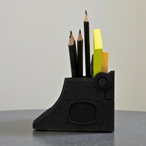 May include: A black desk organizer with a unique, industrial design. It holds several black pencils and yellow and orange sticky notes. The organizer has a curved shape with circular details.