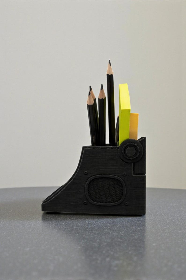May include: A black desk organizer with a unique, industrial design. It holds several black pencils and yellow and orange sticky notes. The organizer has a curved shape with circular details.