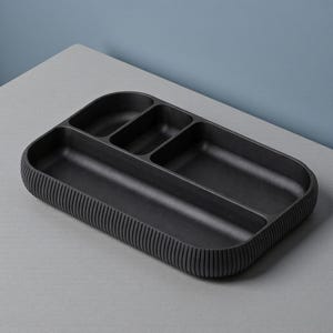 May include: A black rectangular organizer tray with multiple compartments. The tray has a ribbed design along the outer edges and is made of a matte black material. The compartments are various sizes and shapes.