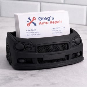 May include: A black car-shaped business card holder with a stack of white cards. The cards have the text "Greg's Auto Repair" and contact information. The holder is made of a matte black material.