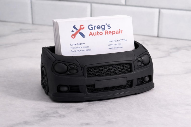 May include: A black car-shaped business card holder with a stack of white cards. The cards have the text "Greg's Auto Repair" and contact information. The holder is made of a matte black material.