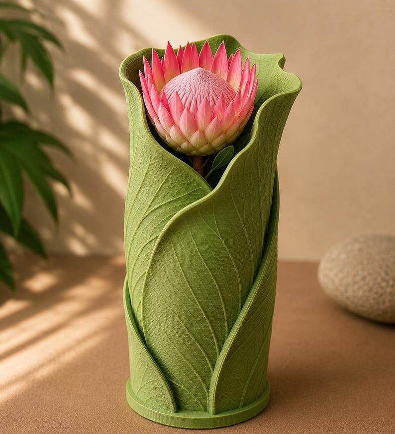 May include: A green vase shaped like overlapping leaves, holding a pink protea flower. The vase has a textured surface and a cylindrical shape. The flower has a spiky, layered appearance with a light pink hue.