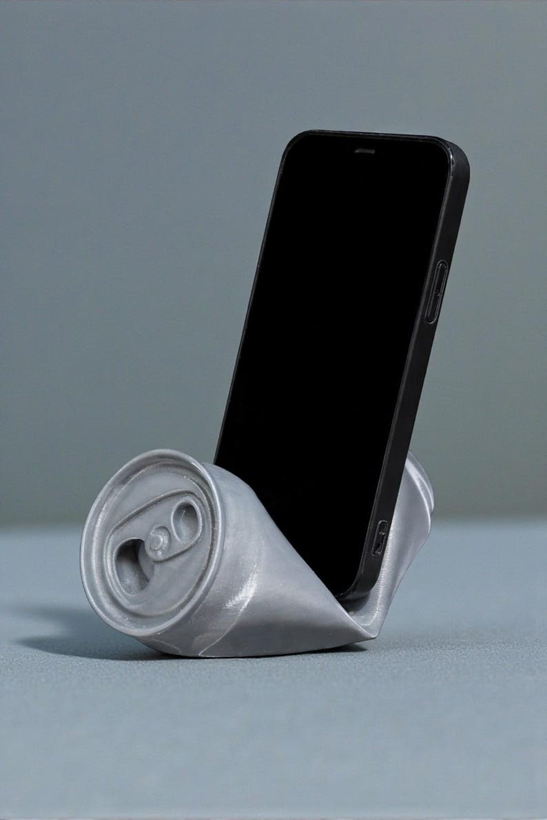 May include: A silver-colored phone stand shaped like a crushed soda can. A black smartphone is placed in the stand, with the screen facing forward. The stand is on a gray surface.