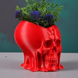 May include: A red skull-shaped planter with a textured surface. The planter is filled with green and purple artificial plants. The skull has detailed facial features and a unique, melted appearance. The planter is a decorative item.