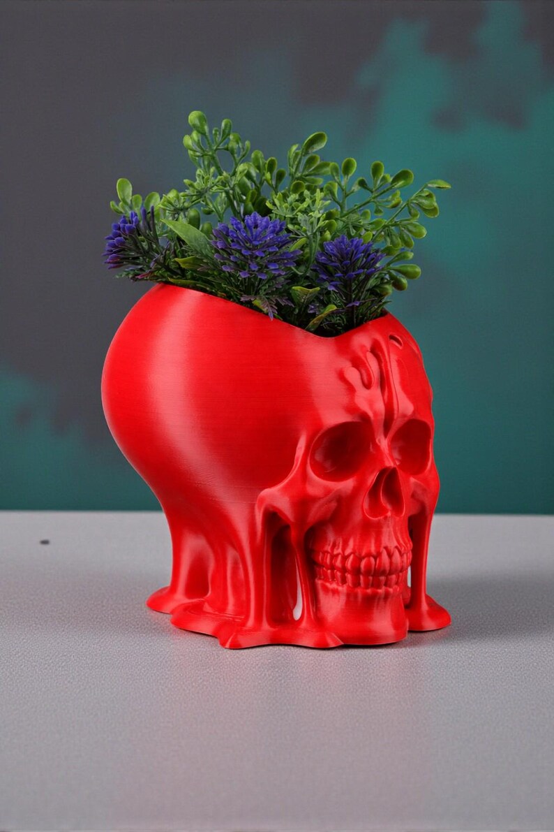 May include: A red skull-shaped planter with a textured surface. The planter is filled with green and purple artificial plants. The skull has detailed facial features and a unique, melted appearance. The planter is a decorative item.