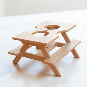 May include: A miniature picnic table in a light brown color. The table has a rectangular top with two circular cutouts and attached benches. The table is approximately 6 inches long, 4 inches wide, and 3 inches tall.