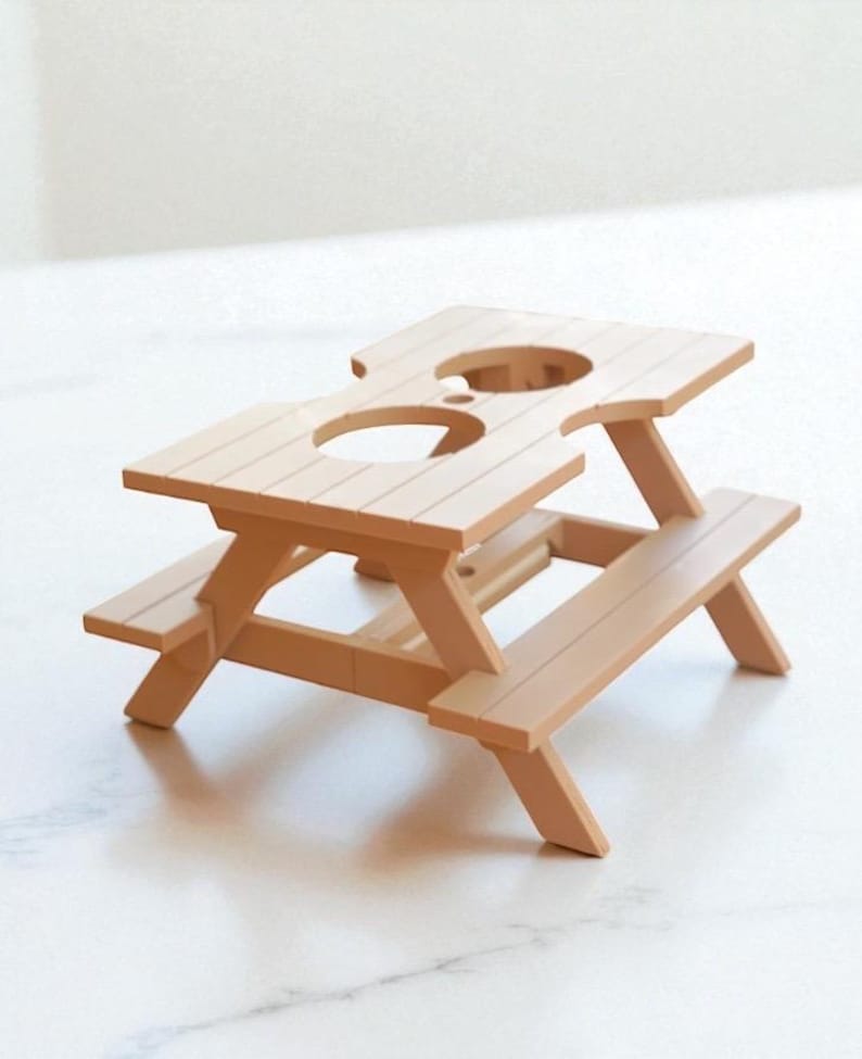 May include: A miniature picnic table in a light brown color. The table has a rectangular top with two circular cutouts and attached benches. The table is approximately 6 inches long, 4 inches wide, and 3 inches tall.