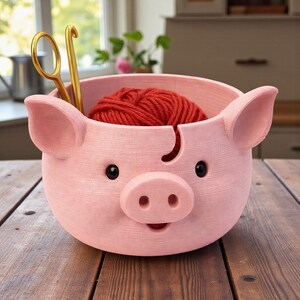 Puede incluir: Un cuenco para hilo en forma de cerdo rosa con ojos negros y un hocico. Una madeja de hilo rojo se encuentra dentro del cuenco, con unas tijeras doradas y un ganchillo. El cuenco tiene una ranura para que pase el hilo.