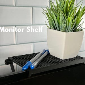 May include: A black monitor shelf with two blue pens on top. The shelf is mounted on a black computer monitor. A white planter with green plants is sitting on the shelf. The text "Monitor Shelf" is visible in the image.