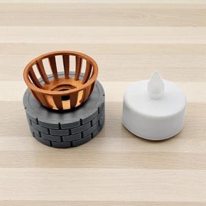 May include: A decorative set featuring a small, round, copper-colored basket with vertical slats, atop a gray, brick-patterned base. A white, battery-operated LED candle with a flame-shaped top is next to the set. The items are designed for home decor.