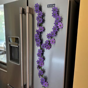 May include: A stainless steel refrigerator with a garland of purple flower magnets. The flowers have green centers and are arranged in a flowing, decorative pattern. The refrigerator has a water dispenser and the Maytag logo.