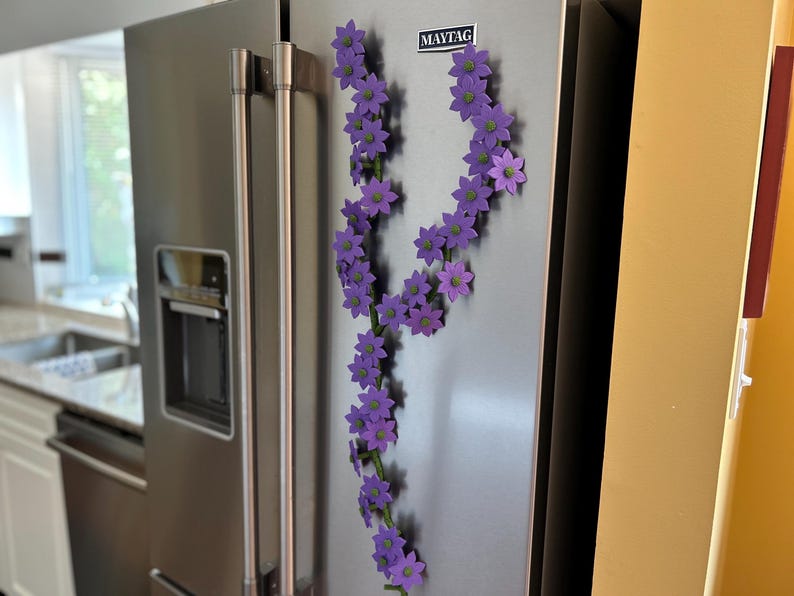 May include: A stainless steel refrigerator with a garland of purple flower magnets. The flowers have green centers and are arranged in a flowing, decorative pattern. The refrigerator has a water dispenser and the Maytag logo.