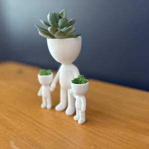May include: A white ceramic planter set in the shape of a family. The adult figure holds the hands of two smaller figures. The heads of each figure are open and filled with green plants. The largest figure has a succulent.