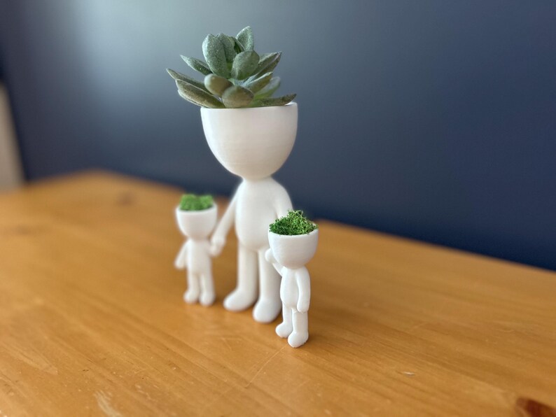May include: A white ceramic planter set in the shape of a family. The adult figure holds the hands of two smaller figures. The heads of each figure are open and filled with green plants. The largest figure has a succulent.