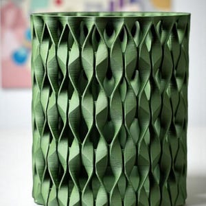 May include: A green cylindrical container with a geometric, woven-like pattern. The container is made of a solid material and has a textured surface. The design features repeating diamond shapes, creating a visually interesting texture. The container is suitable for holding pens, pencils, or other small items.