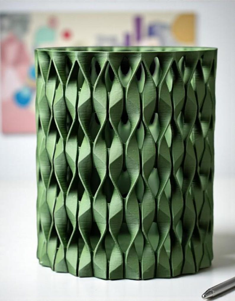 May include: A green cylindrical container with a geometric, woven-like pattern. The container is made of a solid material and has a textured surface. The design features repeating diamond shapes, creating a visually interesting texture. The container is suitable for holding pens, pencils, or other small items.