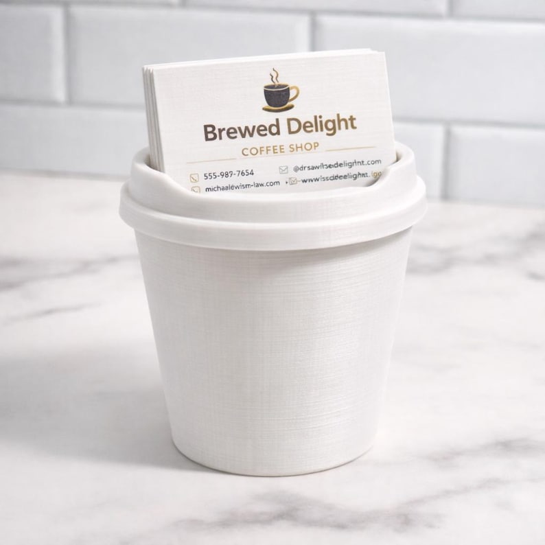 May include: A white, textured coffee cup-shaped business card holder. The holder contains a stack of business cards with the text "Brewed Delight Coffee Shop" and contact information. The cup sits on a white surface.