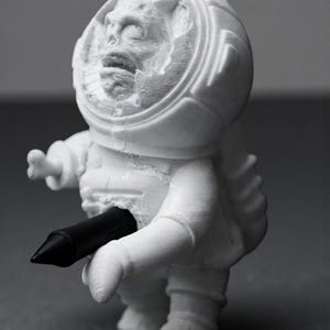 Pu&ograve; includere: Figurina bianca stampata in 3D di un astronauta con un volto dettagliato all'interno del casco. L'astronauta tiene in mano una penna nera. La figurina ha una superficie strutturata.
