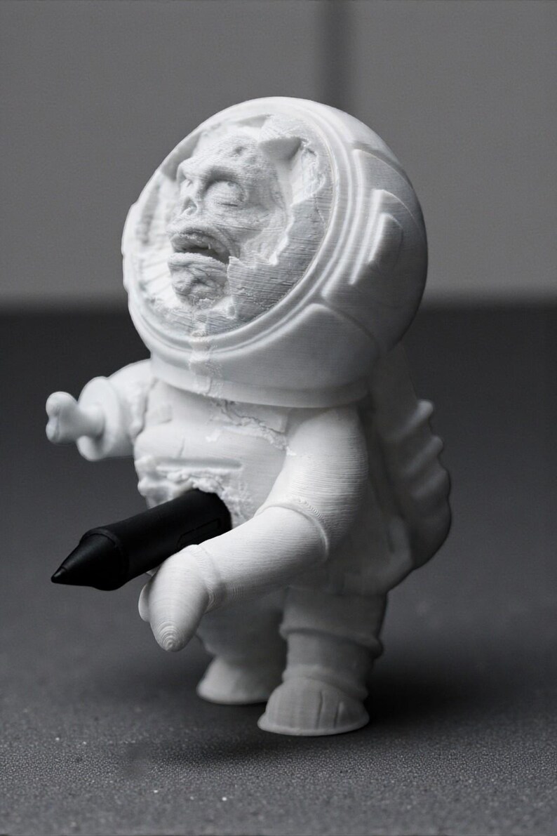 Pu&ograve; includere: Figurina bianca stampata in 3D di un astronauta con un volto dettagliato all'interno del casco. L'astronauta tiene in mano una penna nera. La figurina ha una superficie strutturata.