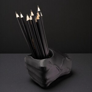 May include: A black pencil holder shaped like a fist, filled with black pencils with white tips. The pencils are neatly arranged inside the holder. The holder is on a dark surface, with a black background.