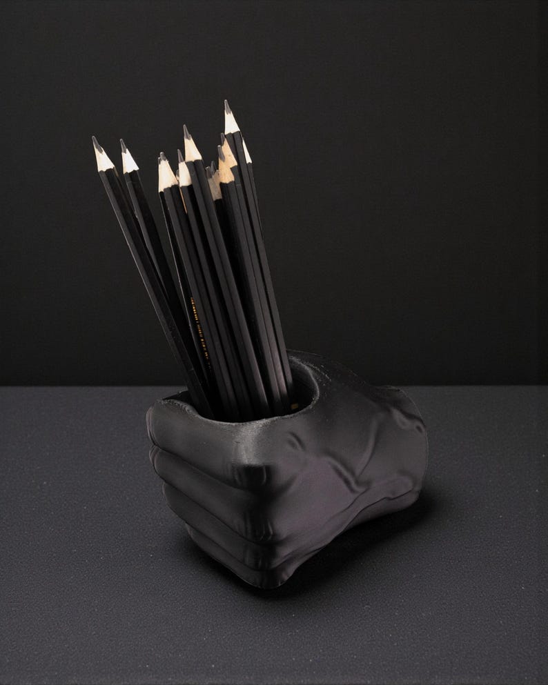 May include: A black pencil holder shaped like a fist, filled with black pencils with white tips. The pencils are neatly arranged inside the holder. The holder is on a dark surface, with a black background.