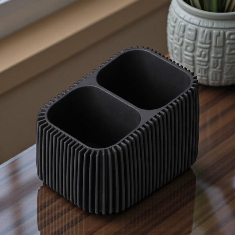 May include: A black desk organizer with two compartments. The organizer has a ribbed exterior design and is made of a matte material. The product is sitting on a reflective surface.