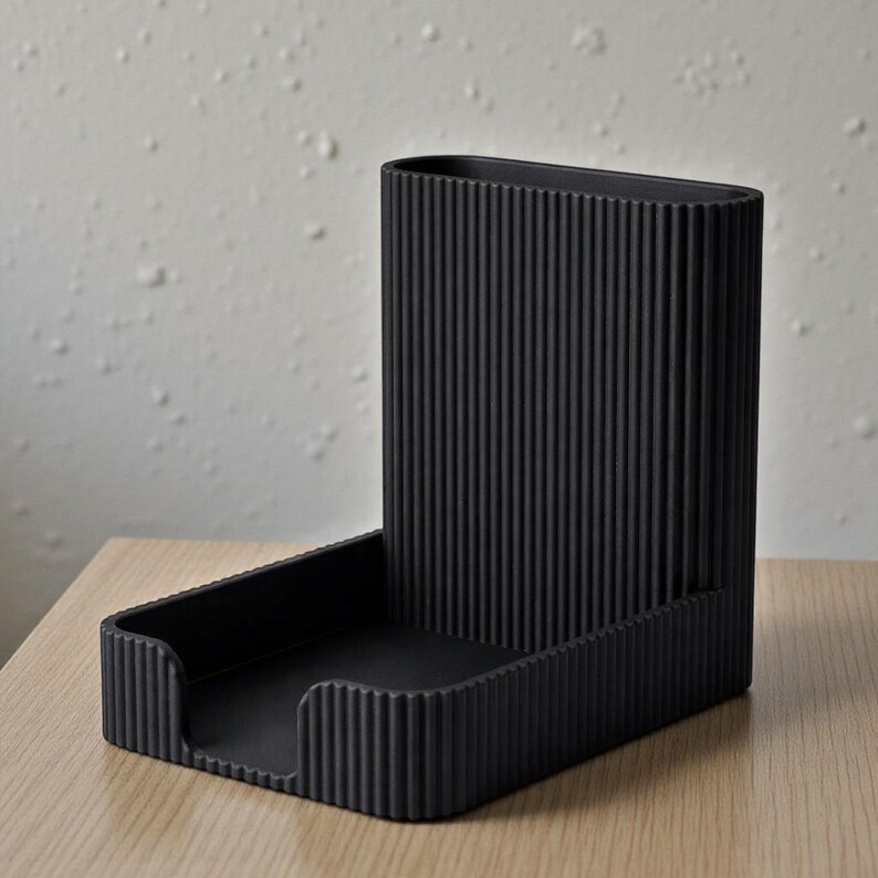 Pu&ograve; includere: Un organizer da scrivania nero con un design a coste verticali. Presenta una sezione alta e rettangolare e un vassoio inferiore rettangolare. Gli articoli sono realizzati in materiale nero opaco e poggiano su una superficie in legno chiaro.