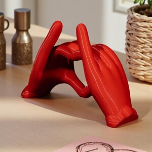 May include: A vibrant red sculpture of two hands forming a heart shape. The glossy finish of the hands contrasts with the neutral background. A small pink notepad with a heart and the word "LOVE" is visible.