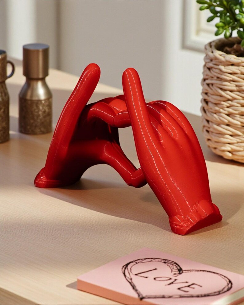 May include: A vibrant red sculpture of two hands forming a heart shape. The glossy finish of the hands contrasts with the neutral background. A small pink notepad with a heart and the word "LOVE" is visible.