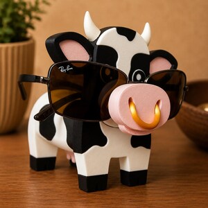 May include: A whimsical cow-shaped eyeglass holder. The holder is painted in black and white with a pink nose and gold ring. The cow is wearing a pair of Ray-Ban sunglasses. The holder is made of wood and is a decorative item.