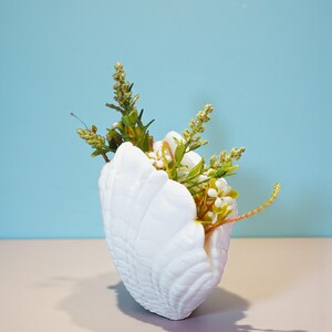 May include: A white seashell-shaped vase filled with an arrangement of artificial flowers and greenery. The vase has a textured surface and sits on a light-colored surface against a blue backdrop. The flowers include white blossoms and various green stems.