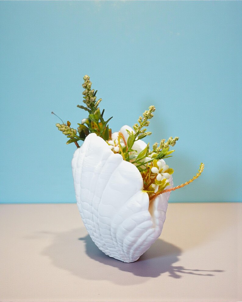 May include: A white seashell-shaped vase filled with an arrangement of artificial flowers and greenery. The vase has a textured surface and sits on a light-colored surface against a blue backdrop. The flowers include white blossoms and various green stems.