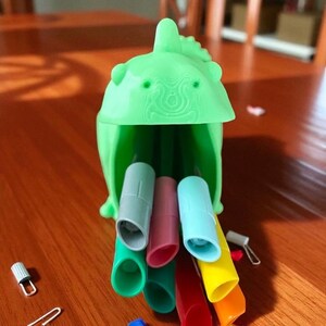 K&ouml;nnte beinhalten: Ein gr&uuml;ner Stifthalter aus Kunststoff in Dinosaurierform mit mehreren bunten Stiftkappen in verschiedenen Farben, darunter Gr&uuml;n, Blau, Rosa und Gelb. Der Stifthalter befindet sich auf einer Holzoberfl&auml;che.