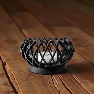 May include: A black, geometric candle holder with a white candle inside. The holder has a round base and an open, lattice-like design. The candle holder is made of metal and sits on a wooden surface.