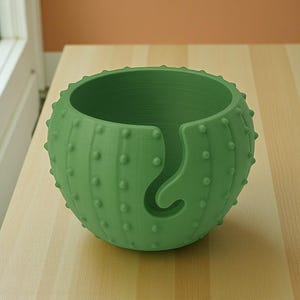 May include: A green cactus-shaped yarn bowl with a curved yarn guide. The bowl has a textured surface with small, rounded bumps, and is sitting on a light-colored wooden surface. The yarn bowl is designed for knitting and crochet projects.
