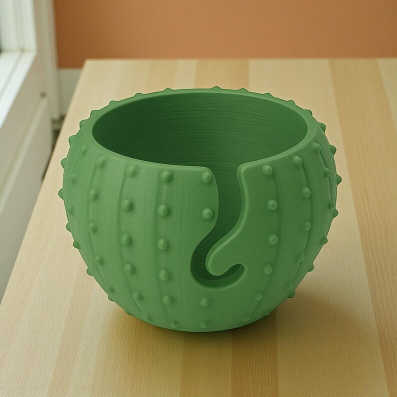 May include: A green cactus-shaped yarn bowl with a curved yarn guide. The bowl has a textured surface with small, rounded bumps, and is sitting on a light-colored wooden surface. The yarn bowl is designed for knitting and crochet projects.