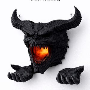 May include: A black wall-mounted sculpture of a demonic face with large horns, sharp teeth, and outstretched hands. The mouth glows with an orange light. The text "mounts to wall with screws (not included)" is above the sculpture.